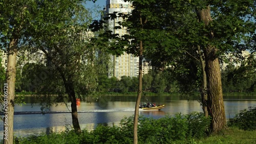A wide shot of a yellow rescue boat speeding on a calm river with passengers on board, framed by green trees with city residential high-rise buildings in the background