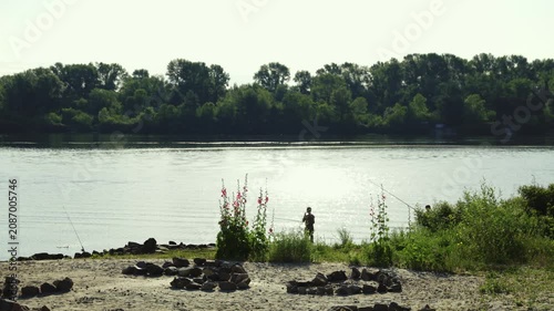 A wide cinematic shot of a lone fisherman silhouette standing on a sandy riverbank with a fishing rod, looking out at the calm water reflecting bright morning sunlight