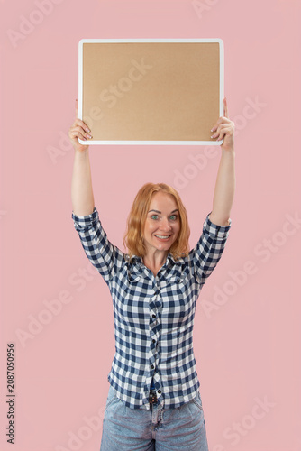 Young woman with red hair and a cheerful expression holding up a blank rectangular sign or board above her head, ready for a personalized message or advertisement on a pink background