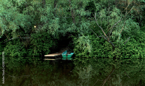 Small rowboat moored beside wooden dock under dense riverside trees with calm reflective water and shaded green bank, with rowboat, dock, riverbank, tree detail