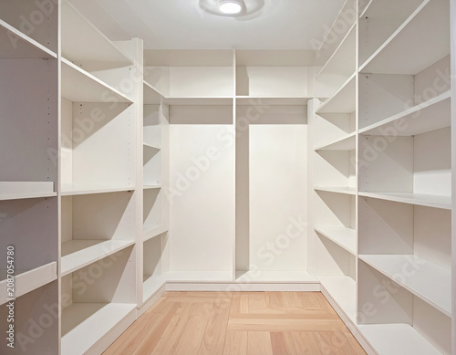 An empty, modern white walk-in closet with built-in shelves and a light wood floor is shown.