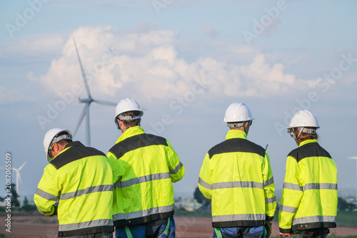 Fotografie Four wind energy engineers in matching yellow watch the rotors churn the evening sky overlooking the turbine studded plain in amber golden hour at a renewable energy site in Thailand