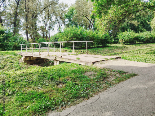 Concrete Footbridge Over Small Creek in Green Park With Trees and Walking Path.