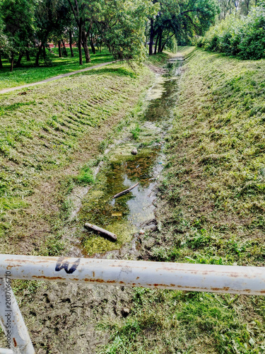 Overgrown Park Drainage Ditch With Shallow Creek, Rusty Railing and Trees in Summer Greenery.