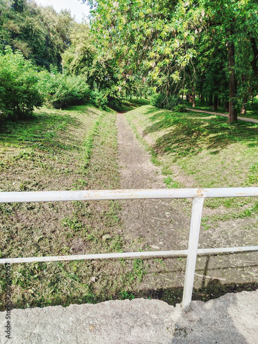 Sunny Green Park Pathway Through Trees With White Metal Railing Over Small Dry Streambed.