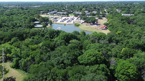 Public gathering grounds sit beyond dense woodland surrounding Leonard and Helen Johns Community Park. Shimmering pond waters mirror vibrant event tents, colorful structures in Flower Mound, TX