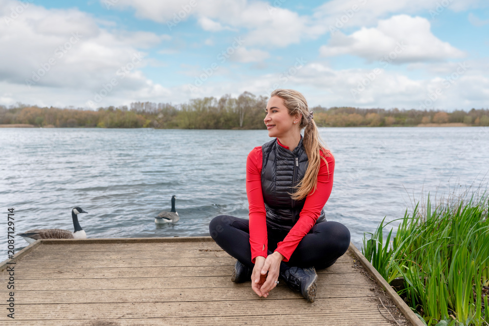 Fototapeta premium Woman sits on dock by water with geese while enjoying the outdoors on a sunny day in a park