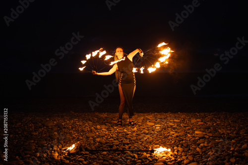Female fire performer spinning flaming fans on a pebble beach at night.