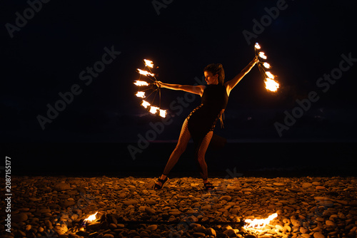 Female fire performer spinning flaming fans on a pebble beach at night.