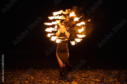 Female fire performer spinning flaming fans on a pebble beach at night.