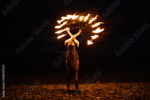 Female fire performer spinning flaming fans on a pebble beach at night.