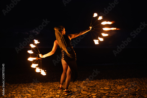 Female fire performer spinning flaming fans on a pebble beach at night.
