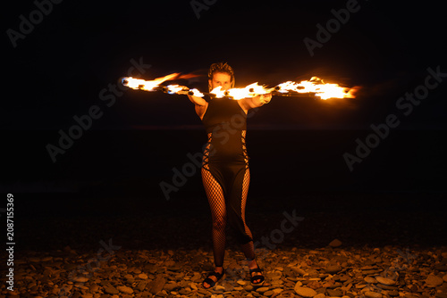 Female fire performer spinning flaming fans on a pebble beach at night.
