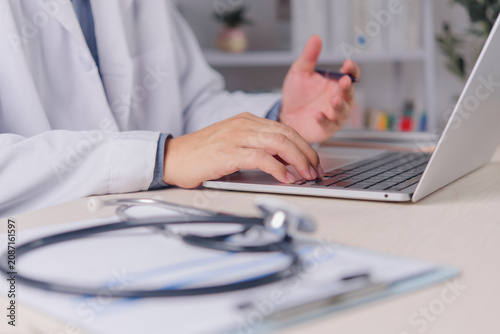 male doctor physician white coat typing laptop computer keyboard desk stethoscope clipboard foreground hospital clinic healthcare professional online consultation electronic medical record working