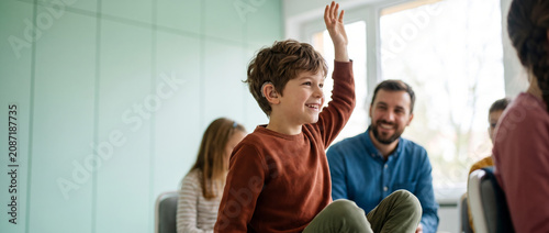 Boy with a hearing aid participating in a classroom discussion. Family support group or teacher visible in the background. Concept of pediatric hearing care and inclusive learning.
