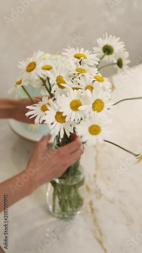 Female hands arranging a fresh chamomile bouquet in a glass vase next to a celebratory cake
