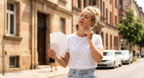 A woman sweating and trying to cool off with a white hand fan on a hot summer day in a city street.