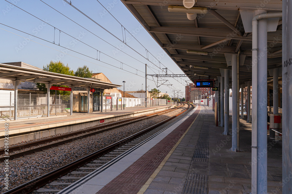 Obraz premium A suburban commuter rail station in Valencia, Spain, featuring covered platforms, overhead electric wires, digital departure boards, and passengers waiting under warm evening sunlight.