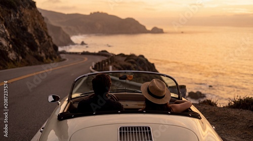 Obraz na płótnie Couple driving convertible car on scenic coastal road at sunset