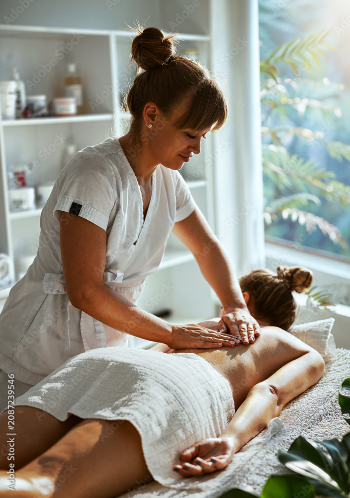 Naklejka premium A mature female therapist concentrates, as she presses both hands firmly onto a young womans upper back during a professional massage session bathed in warm window light.