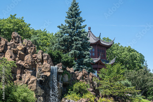 Traditional Asian pavilion surrounded by green trees and pines, situated behind a rocky hill with a small cascading waterfall under a clear blue sky.