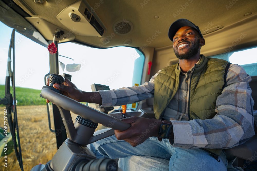 Fototapeta premium A farmer is seen smiling while driving a tractor across a field. The sun is shining, and crops are growing in the background. The scene shows the joy of farming and hard work.