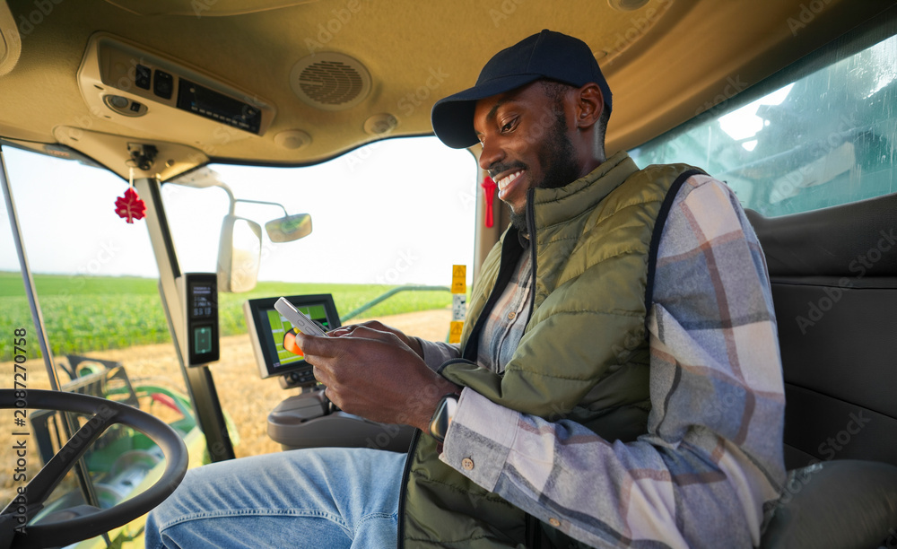 Fototapeta premium Afro American farmer checking information on a smartphone while operating harvesting equipment in a wheat field.