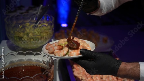 A professional caterer wearing black gloves serves a gourmet plate of shrimp cocktail with avocado and sauce at an upscale event.