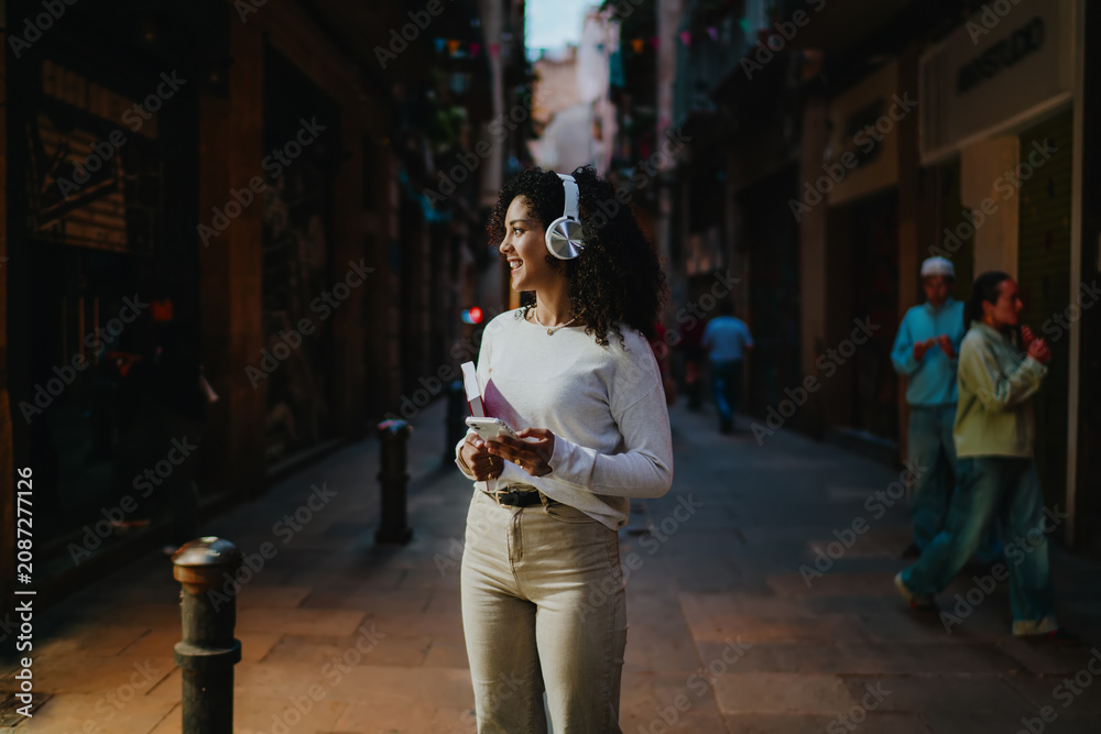 Fototapeta premium Happy young woman in white wireless headphones listening to music and holding smartphone while walking on cozy city street, cheerful female student enjoying audio podcast connection