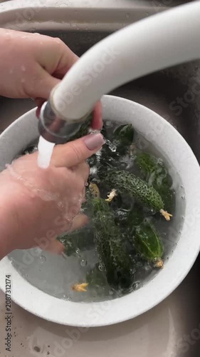 A close-up shot of hands thoroughly washing freshly harvested green cucumbers in a bowl under running tap water, focusing on food hygiene, preparation, and healthy organic lifestyle.