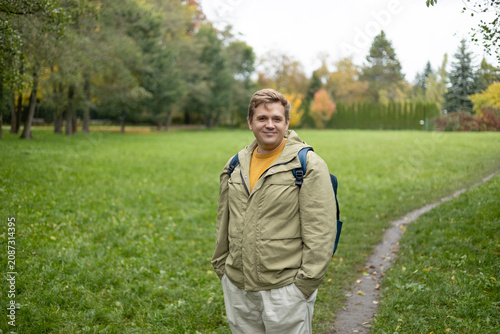 Happy 30s blonde man with backpack standing in the countryside. Traveler. Autumn time. Positive handsome blonde male standing outdoor