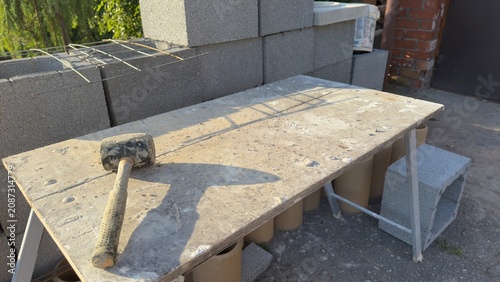 Concrete blocks stacked for construction. Hammer rests on work surface. Poured concrete forms base. Metal supports hold structure upright. Outdoor building site under sun. Ideal for construction, DIY