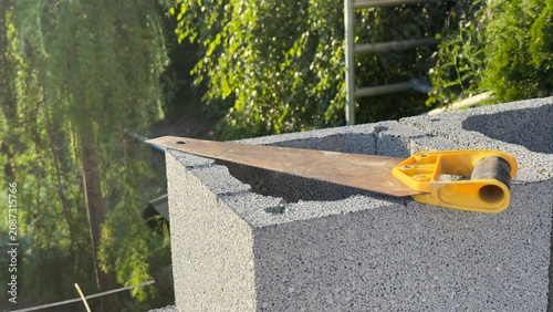 Yellow-handled hand saw rests on gray concrete blocks. Saw blade shows signs of use. Sunlight highlights texture of bricks and tool. Ideal for construction, DIY, or renovation projects