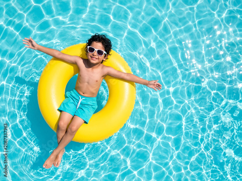 cute indian boy floating on inflatable swim ring in crystal clear swimming pool for summer holiday concept