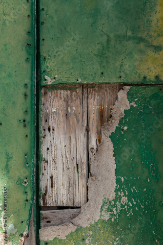 A weathered green metal panel reveals crumbling plaster and rotting wood beneath, showing layers of decay and age