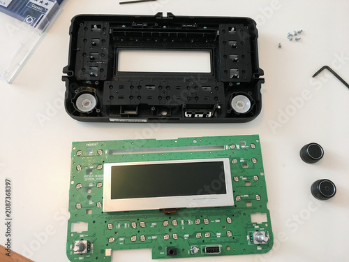 Disassembled car radio with front faceplate and circuit board with display on a table.