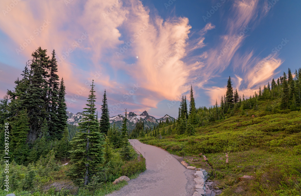 Naklejka premium Paved trail through mountain forest with dramatic sunset clouds