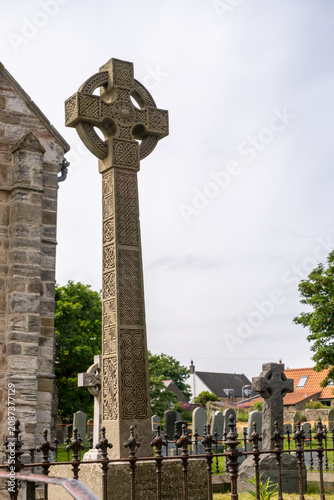 A close, selective-focus view of an ornate Celtic cross in a churchyard, showcasing its intricate stone carvings and craftsmanship against a softly blurred background.