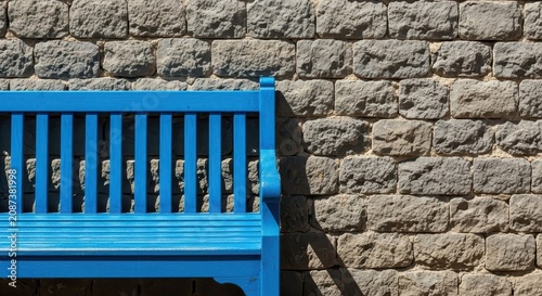 A sturdy empty blue wooden slat bench rests against a rough gray ancient stacked stone wall on a bright sunny day, suggesting rest and quiet, slats, durable, nobody
