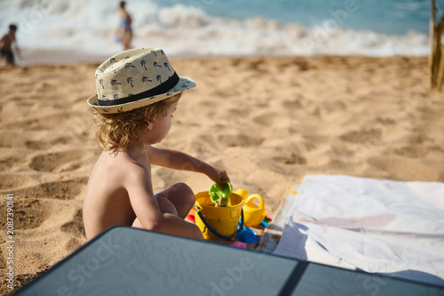 Toddler in a straw hat plays with bucket and shovel on warm sand, enjoying a sunny summer day by the sea with gentle waves and a relaxed family beach atmosphere.