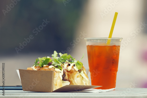 Loaded street food sandwich with fresh herbs and iced tea in a plastic cup, captured at a Karlin food festival in Prague, blurred background provides copy space for text or branding. 
