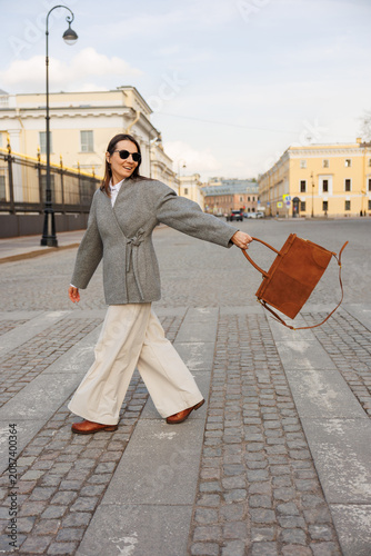 Stylish woman striding across paved city square, carries brown suede bag while wearing sunglasses. Yellow, white classical buildings line background. Confident, sophisticated urban lifestyle moment.