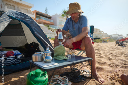 Photography A man sits by a tent on a sandy beach preparing coffee at a low table, surrounded by camping gear and flip-flops, enjoying a relaxed summer seaside morning near coastal houses