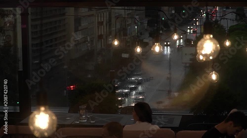 People dine at tables on the rooftop terrace of a cafe during a summer evening rain, the headlights of passing cars shining below, and loft-style interior lamps swaying in the wind from the ceiling.