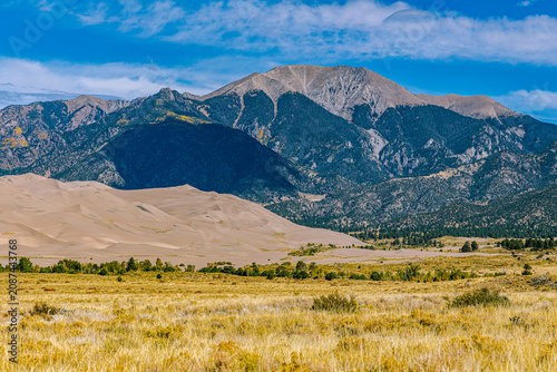Sand dunes nestle below Mount Herard and the Sangre de Cristo range across golden San Luis Valley grassland, Colorado.