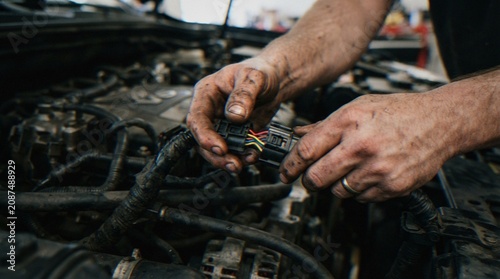 Photos Close-up of a mechanic's greasy hands connecting electrical wiring harness connectors during a car engine repair in a workshop