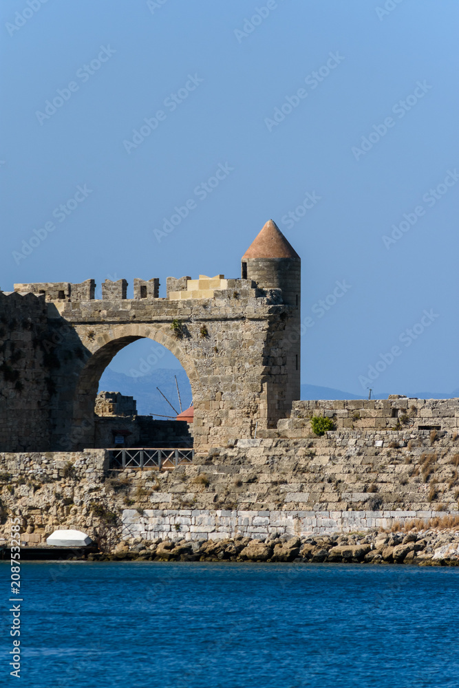 Naklejka premium Detalle de una antigua fortaleza de piedra junto al mar. Muestra un arco, una torre de vigilancia con techo cónico y aguas cristalinas en un día soleado. Ideal para turismo histórico.
