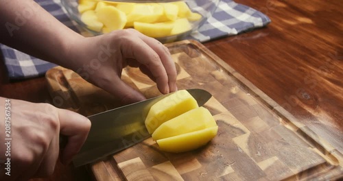Close up of chef's hands cutting a peeled potato into wedges on a wooden cutting board. Fresh potato wedges are being prepared for roasting in the oven as part of a homemade meal.