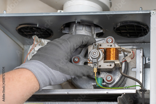 Close-up of a technician hand in a protective glove inspecting an exhaust fan motor and copper induction coil inside an open domestic gas boiler during maintenance.
