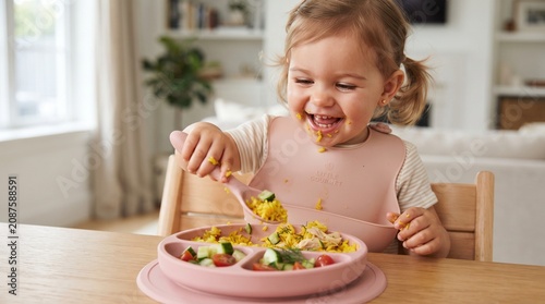 Commercial portrait lifestyle photography of a happy, blue-eyed toddler girl wearing a pink silicone pocket bib during lunchtime – premium baby products branding, healthy children's nutrition marketin